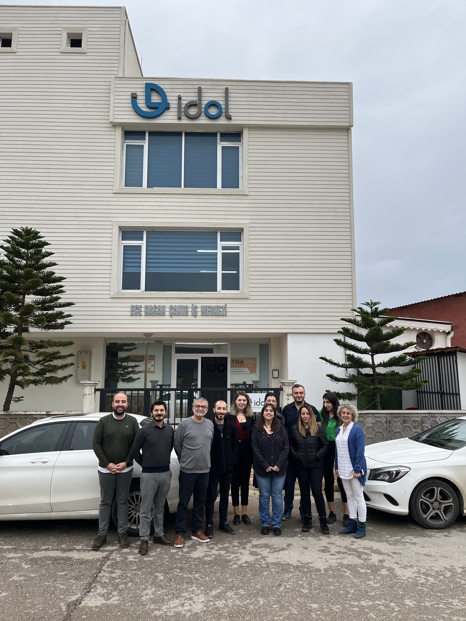 Group of employees posing in front of a beige office building with an 'idol' sign and blue logo, two white cars on either side.